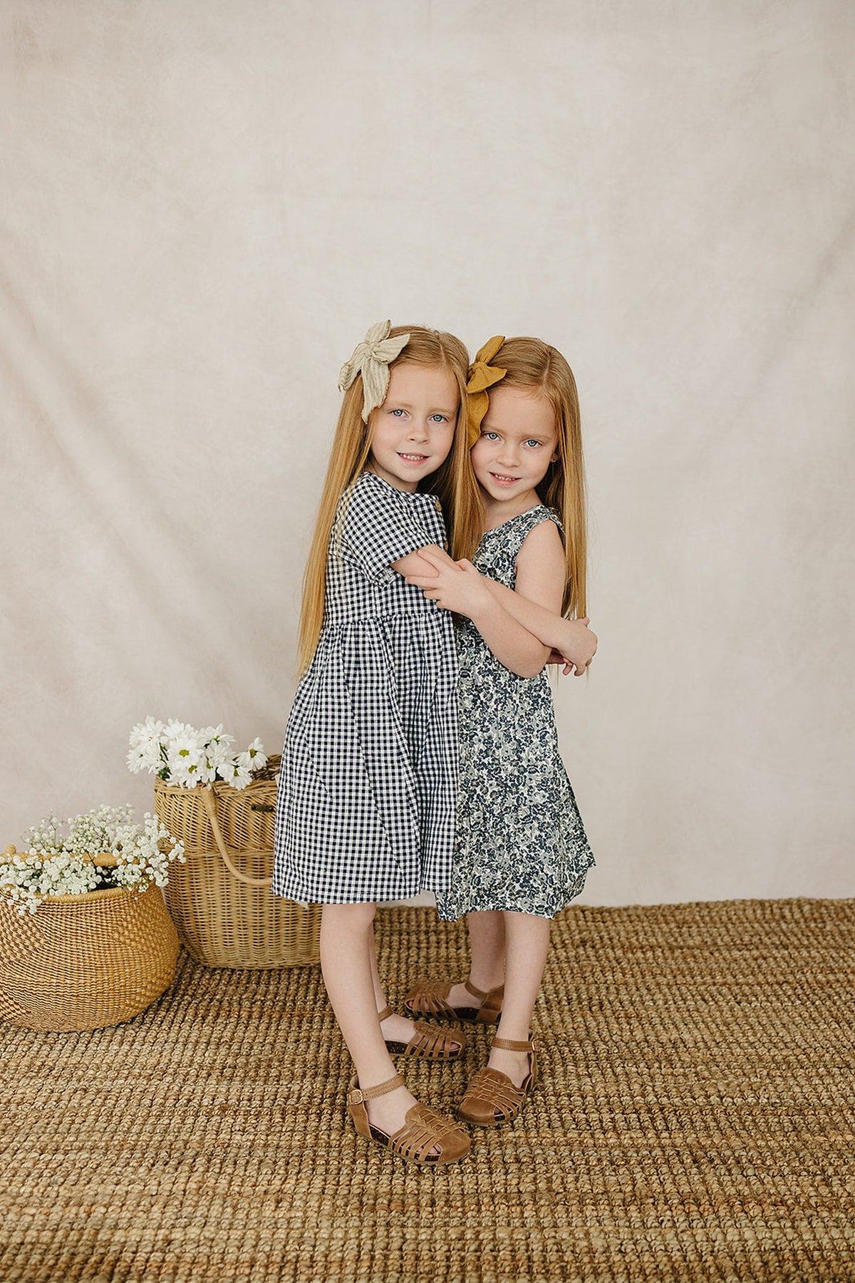 Two young girls embrace, wearing a navy gingham dress and a floral dress, with long hair and matching hair bows.