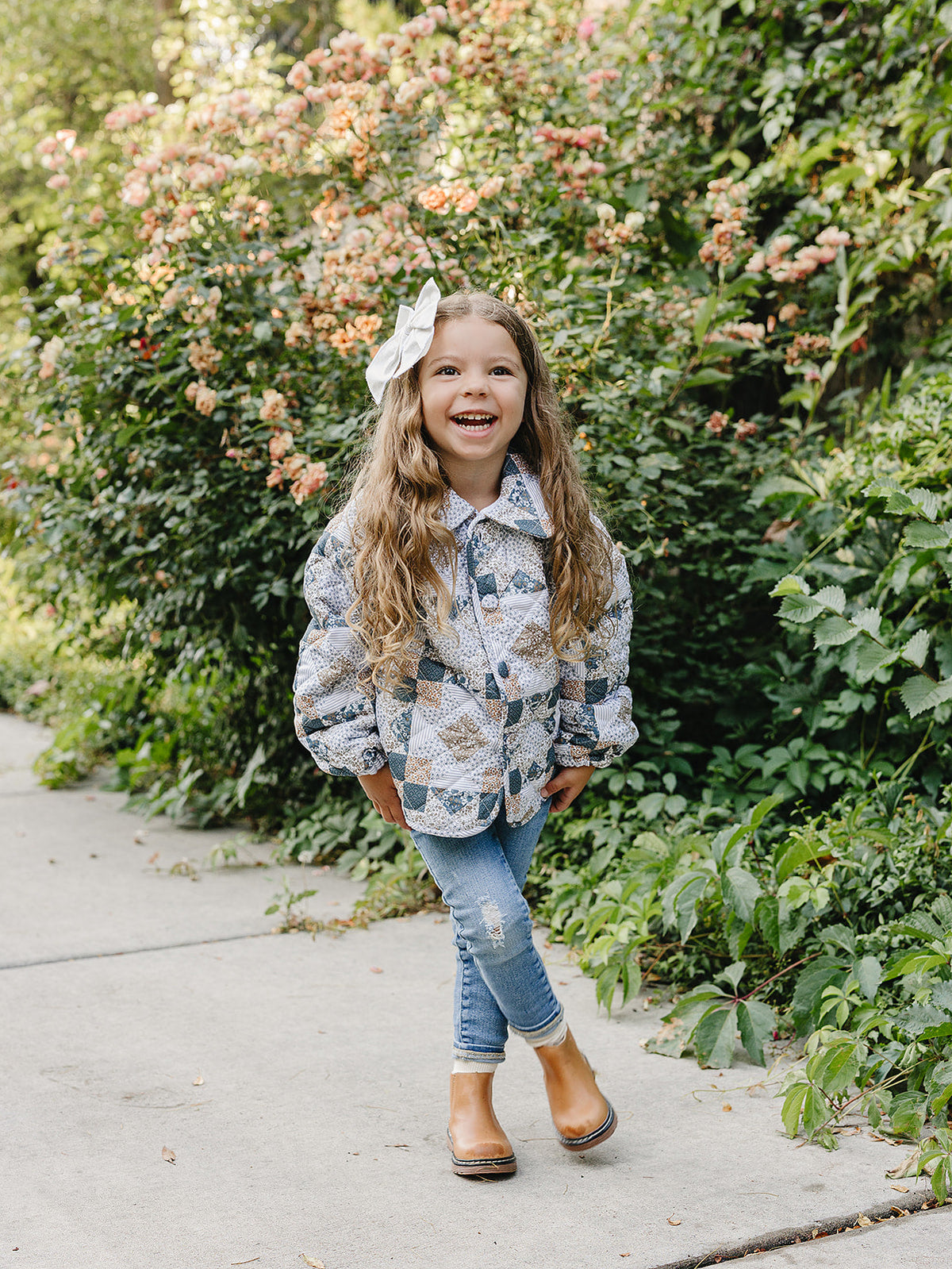 A young girl with long, curly hair smiles while wearing a patchwork quilted cotton jacket, paired with jeans and boots, standing amidst vibrant flowers in a garden setting.
