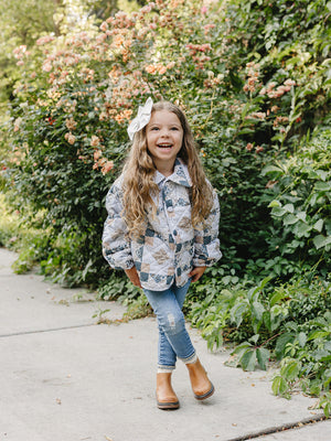 A young girl with long, curly hair smiles while wearing a patchwork quilted cotton jacket, paired with jeans and boots, standing amidst vibrant flowers in a garden setting.