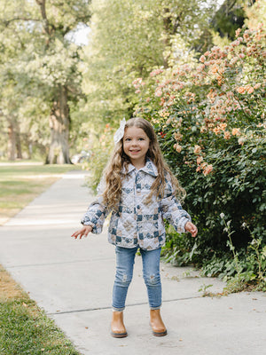A young girl smiles while wearing a patchwork quilted cotton jacket, paired with blue jeans and brown boots, standing on a sidewalk surrounded by blooming flowers and greenery.