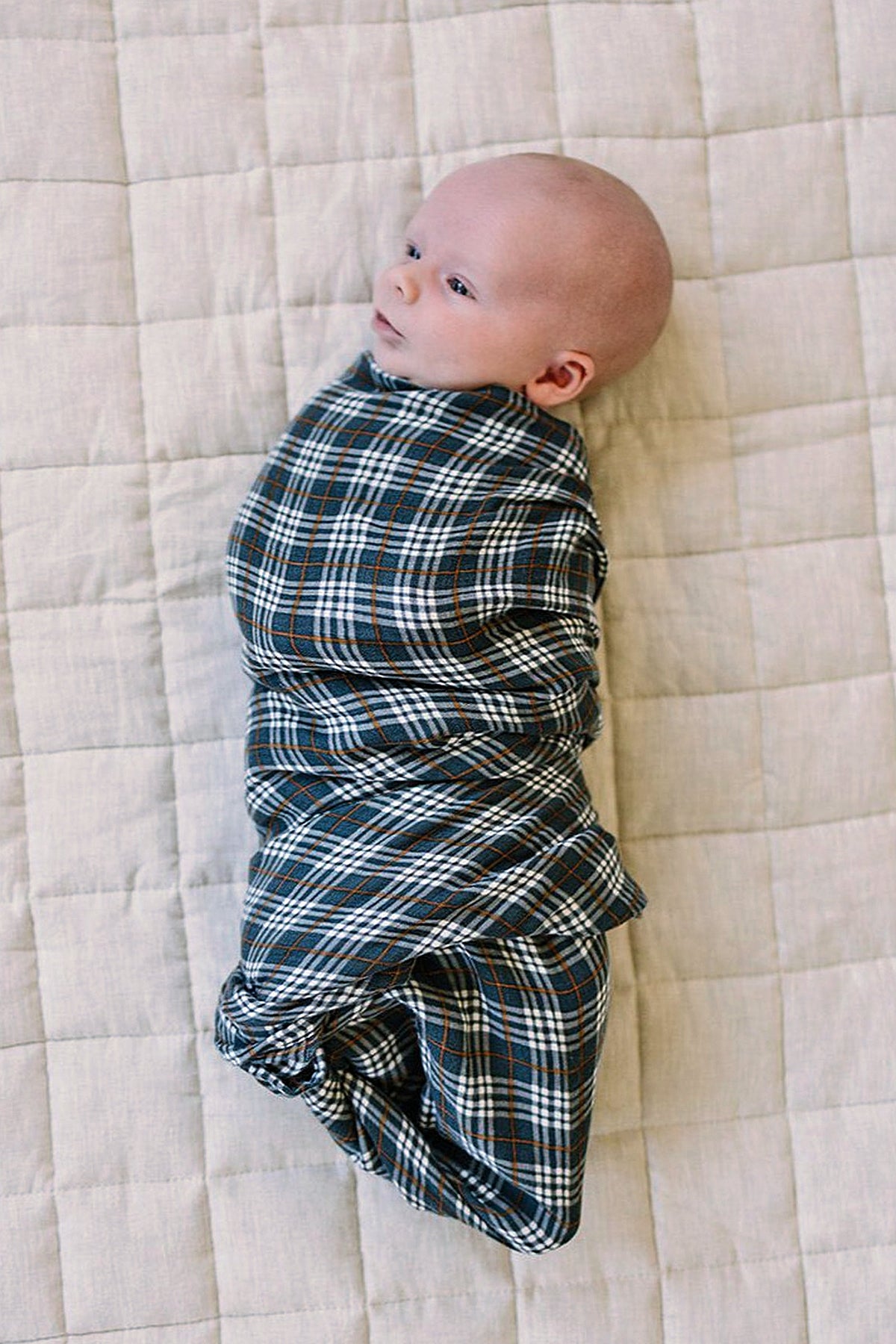 A baby is swaddled in a navy plaid muslin blanket, lying on a light quilted surface.