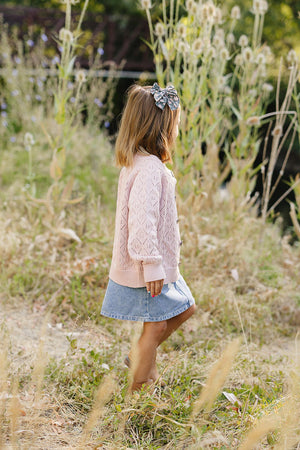 A child wearing a pink pointelle cardigan and denim skirt walks through a grassy field, adorned with a hair bow.