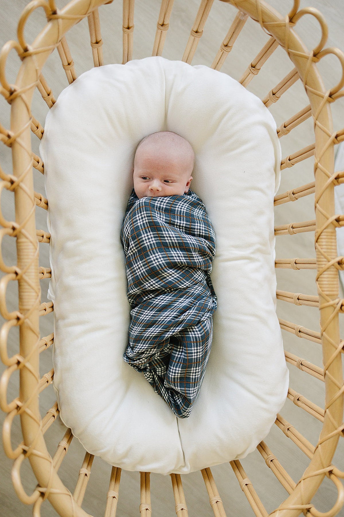 A baby is swaddled in a navy plaid muslin blanket, lying in a cushioned wicker bassinet.