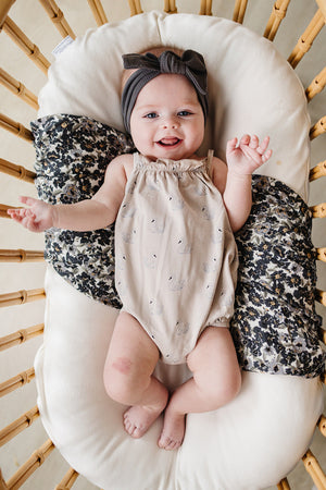 A smiling baby girl wears a cream swan-printed bubble romper, resting on a soft cushion with patterned pillows.