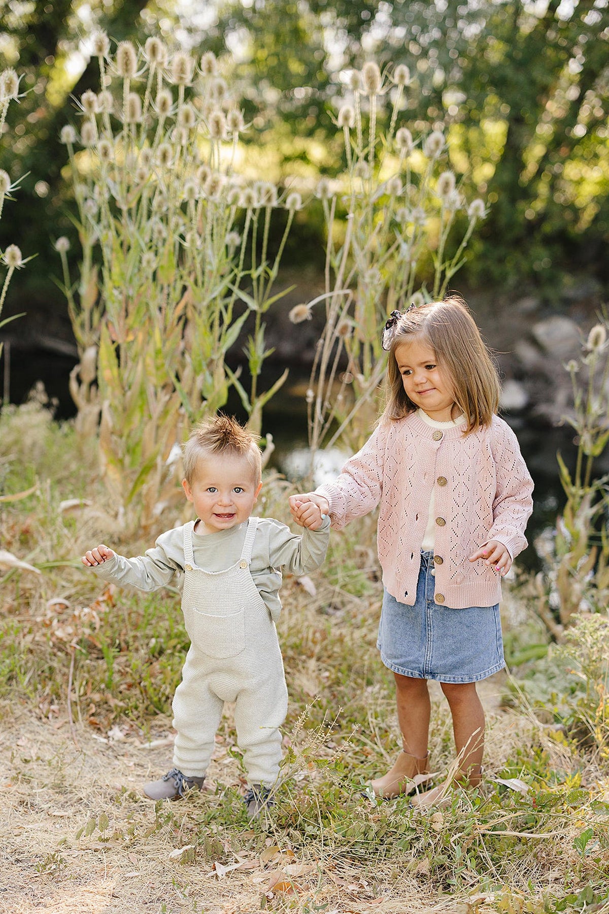 Two children outdoors, one in grey knit overalls and the other in a pink cardigan, holding hands.