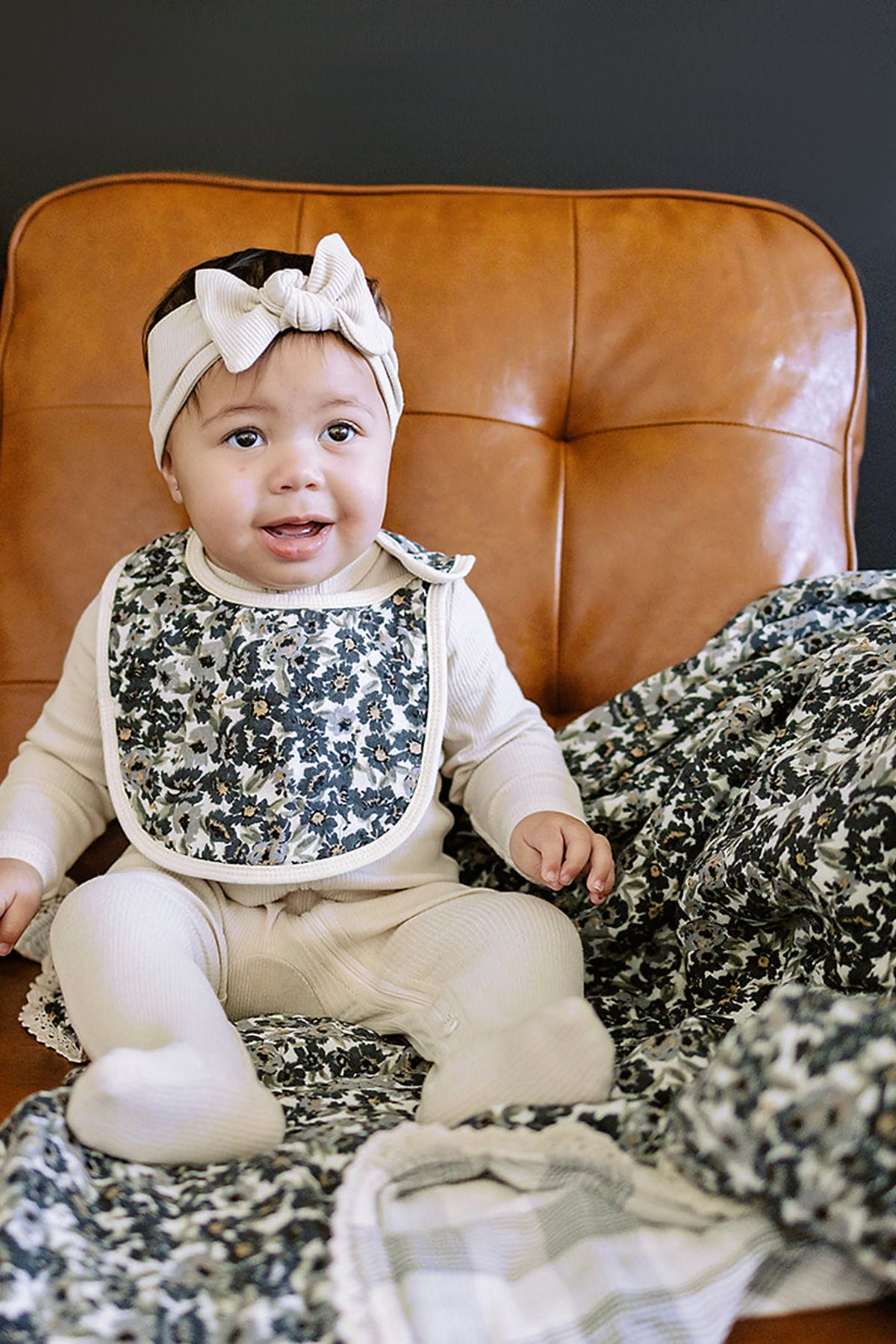 A baby in a beige outfit and floral bib sits on a brown chair with a floral quilt.