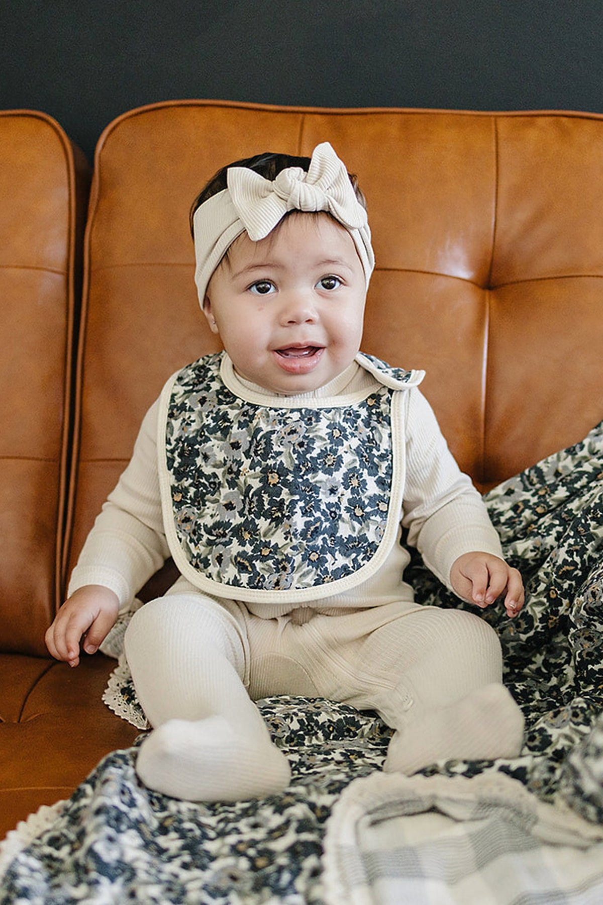 A baby wearing a floral muslin bib and beige outfit sits on a brown couch, smiling.