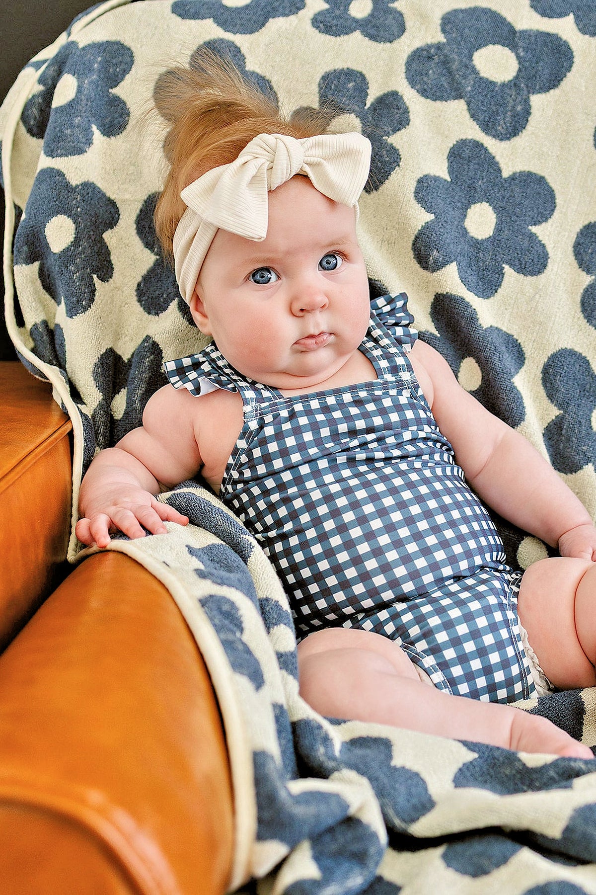 A baby wearing a checkered outfit and bow headband sits on a floral-patterned blanket draped over a chair.