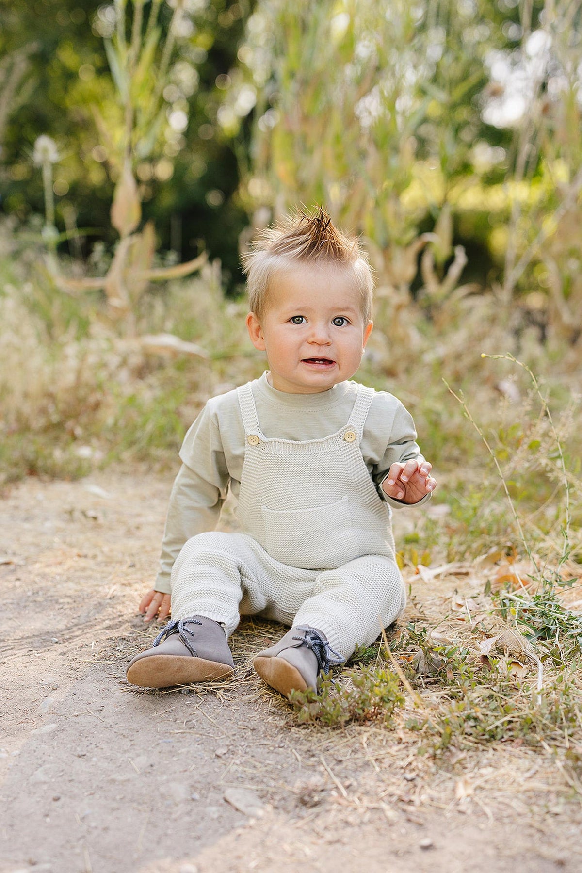 A baby in grey knit overalls sits outdoors on a dirt path, surrounded by greenery.