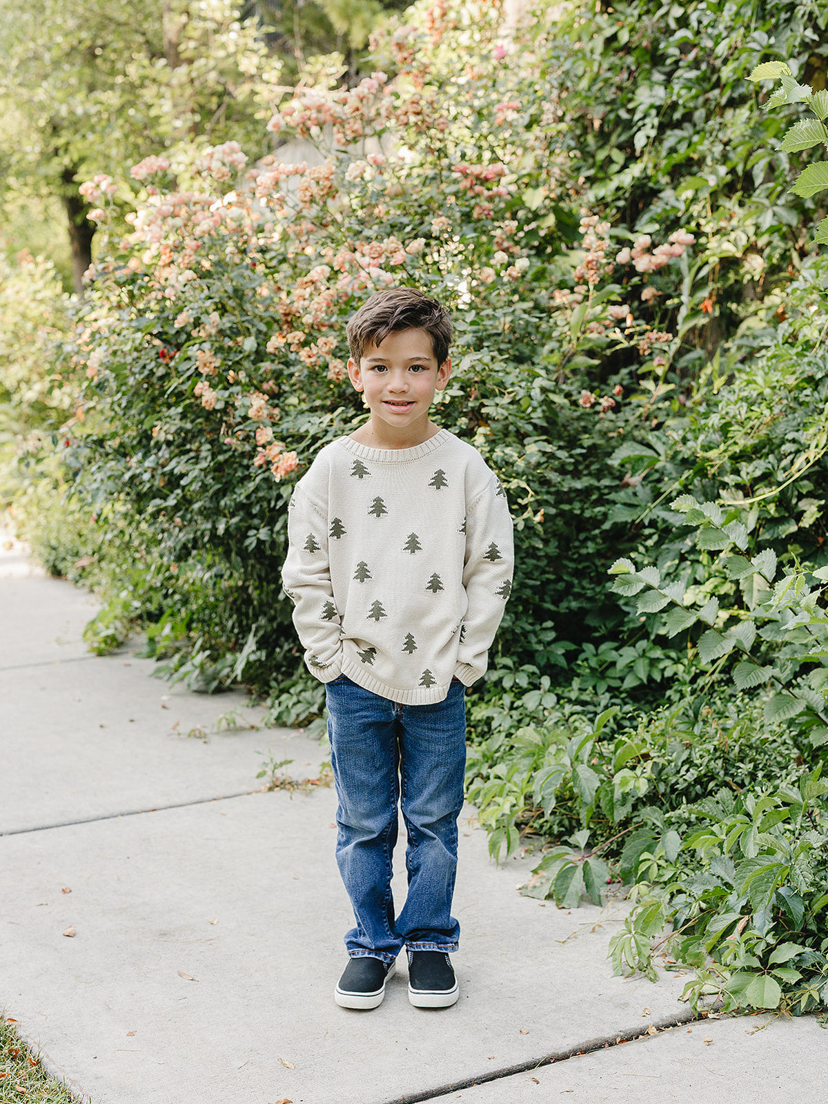 A young boy stands on a sidewalk surrounded by vibrant greenery and blooming flowers. He wears a cozy, ivory knit sweater featuring charming pine tree accents, paired with blue jeans.