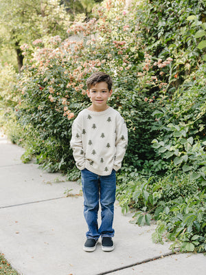 A young boy stands on a sidewalk surrounded by vibrant greenery and blooming flowers. He wears a cozy, ivory knit sweater featuring charming pine tree accents, paired with blue jeans.