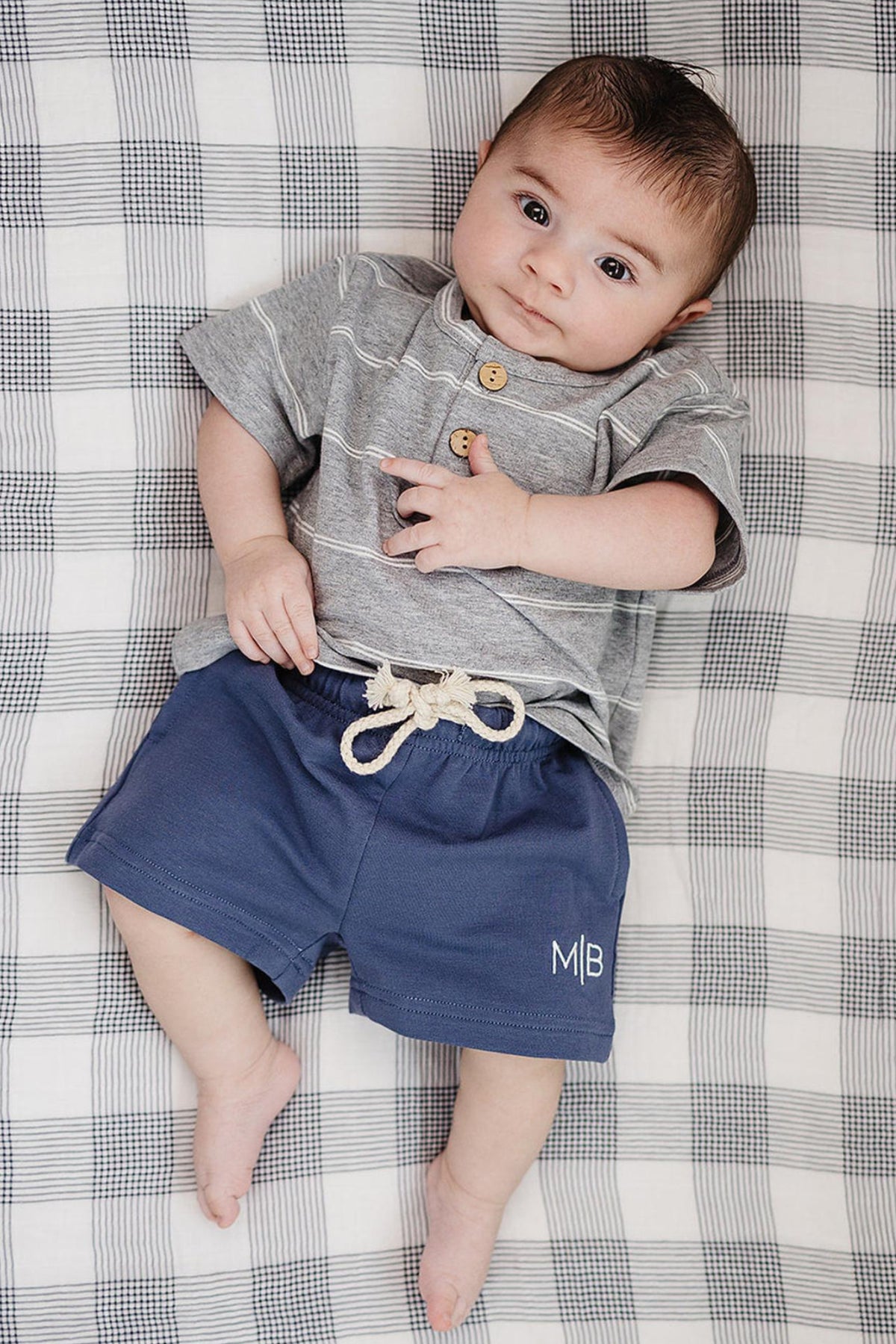 A baby lies on a checkered blanket, wearing a grey striped button-up shirt and navy shorts with a drawstring.