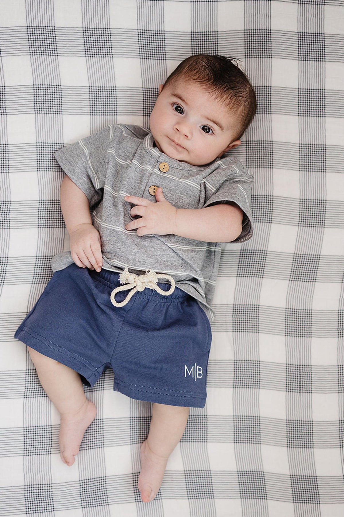A baby lies on a checkered blanket, wearing a gray striped shirt and navy cotton shorts with a drawstring. The relaxed outfit is perfect for comfort and play.