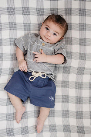 A baby lies on a checkered blanket, wearing a gray striped shirt and navy cotton shorts with a drawstring. The relaxed outfit is perfect for comfort and play.