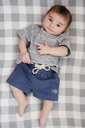 A baby lies on a checkered blanket, wearing a grey striped button-up shirt and navy shorts with a drawstring.