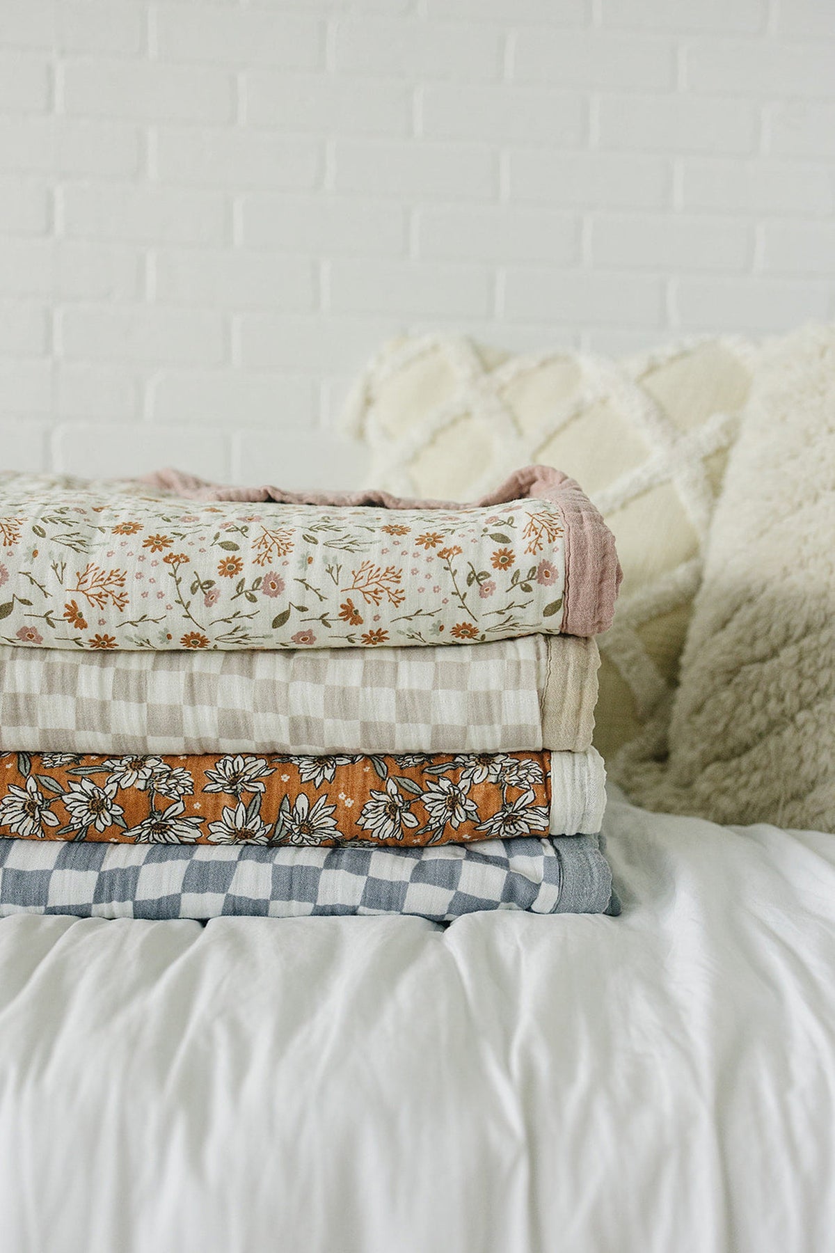 A neatly stacked assortment of muslin quilts featuring floral and checked patterns in soft, muted colors against a white backdrop.
