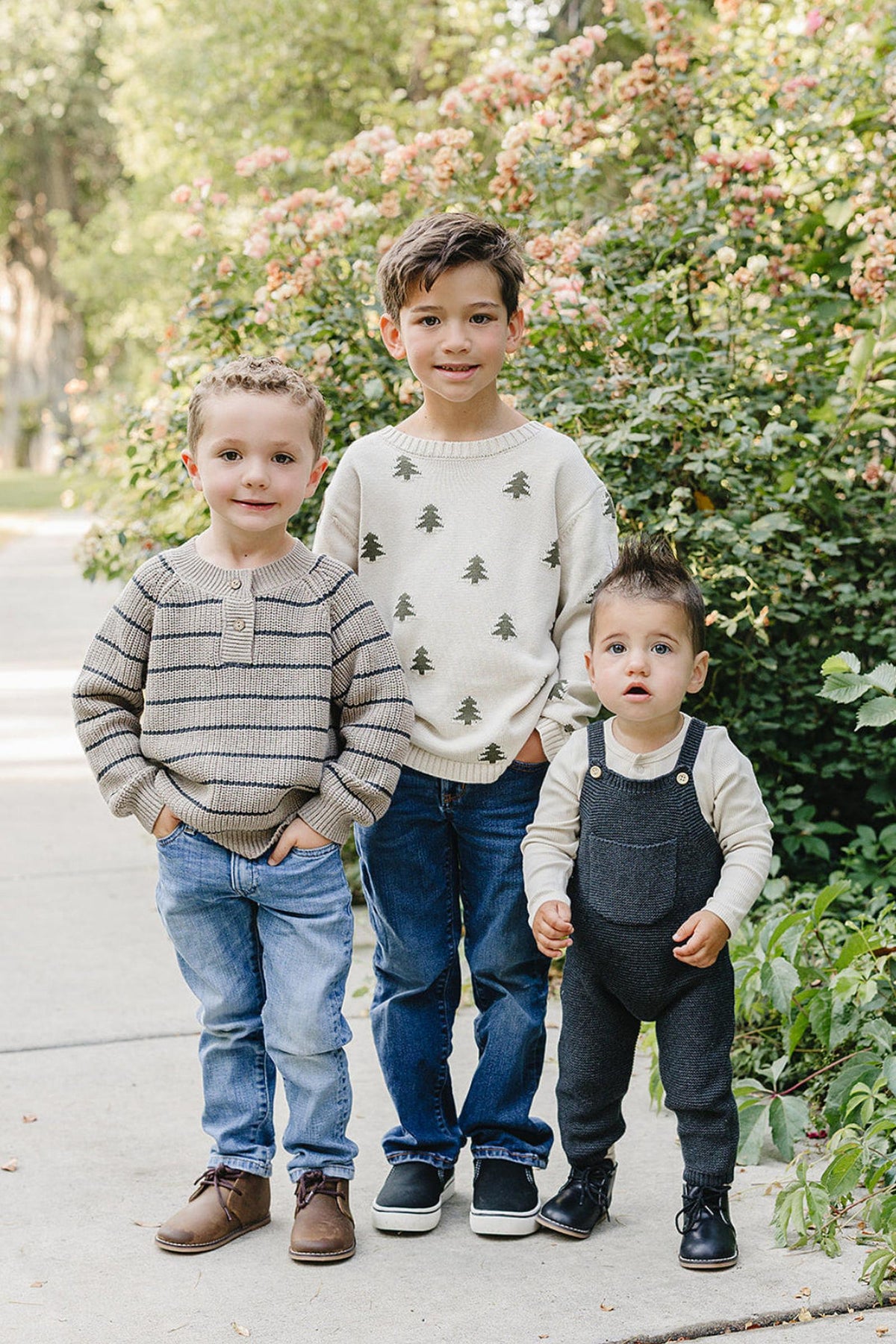 Three young boys stand outdoors on a pathway lined with greenery and flowers. The eldest wears a striped sweater, while the others sport a patterned sweater and overalls, respectively.