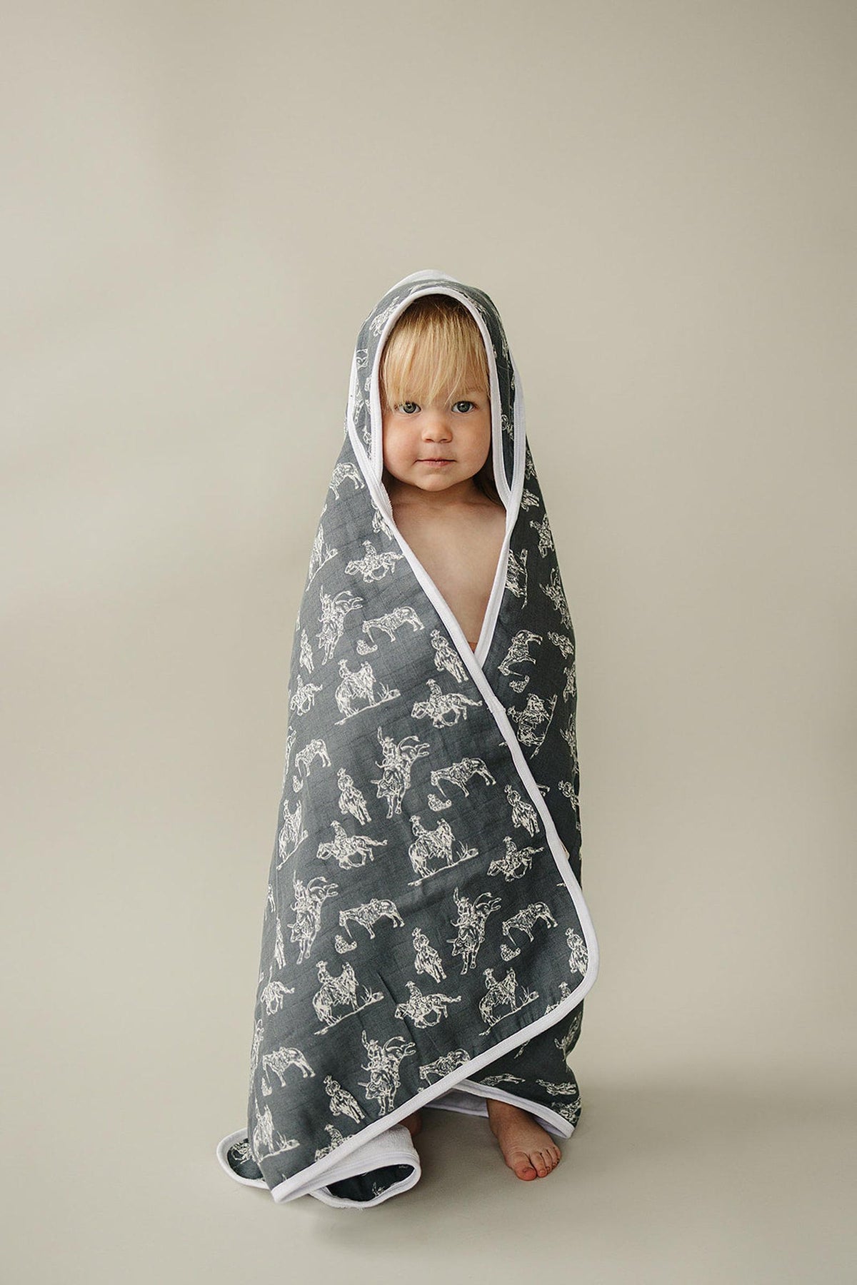 A toddler wrapped in a gray hooded towel with white cowboy patterns and a white trim.
