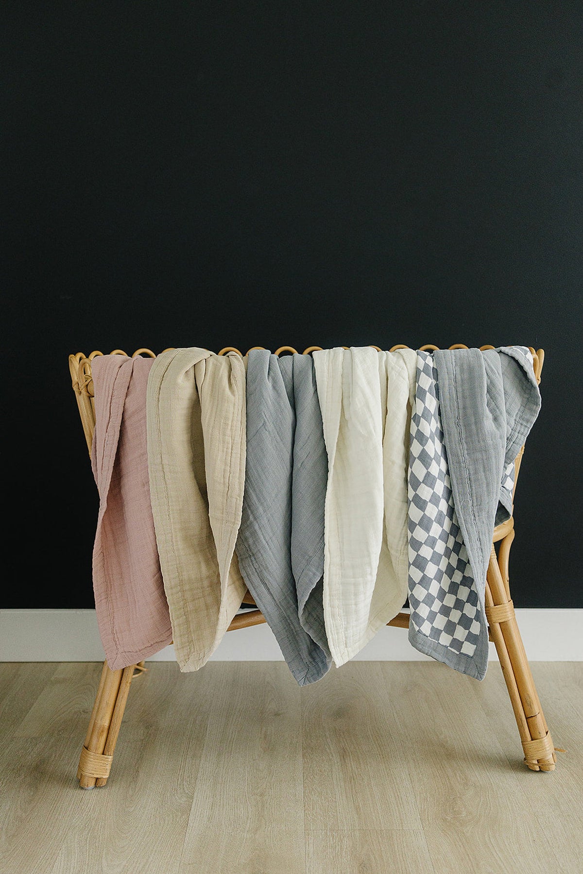 A wicker basket holds six muslin quilts in various colors, including pink, oatmeal, gray, and checkered patterns.
