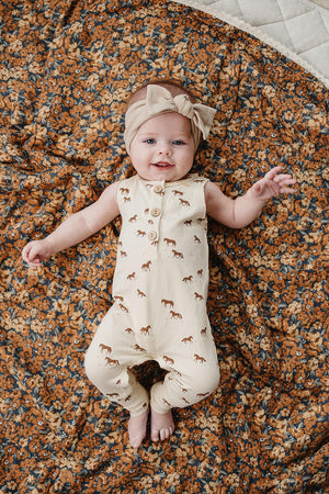 A smiling baby girl wears a cream tank romper with horse graphics, lying on a floral patterned blanket.