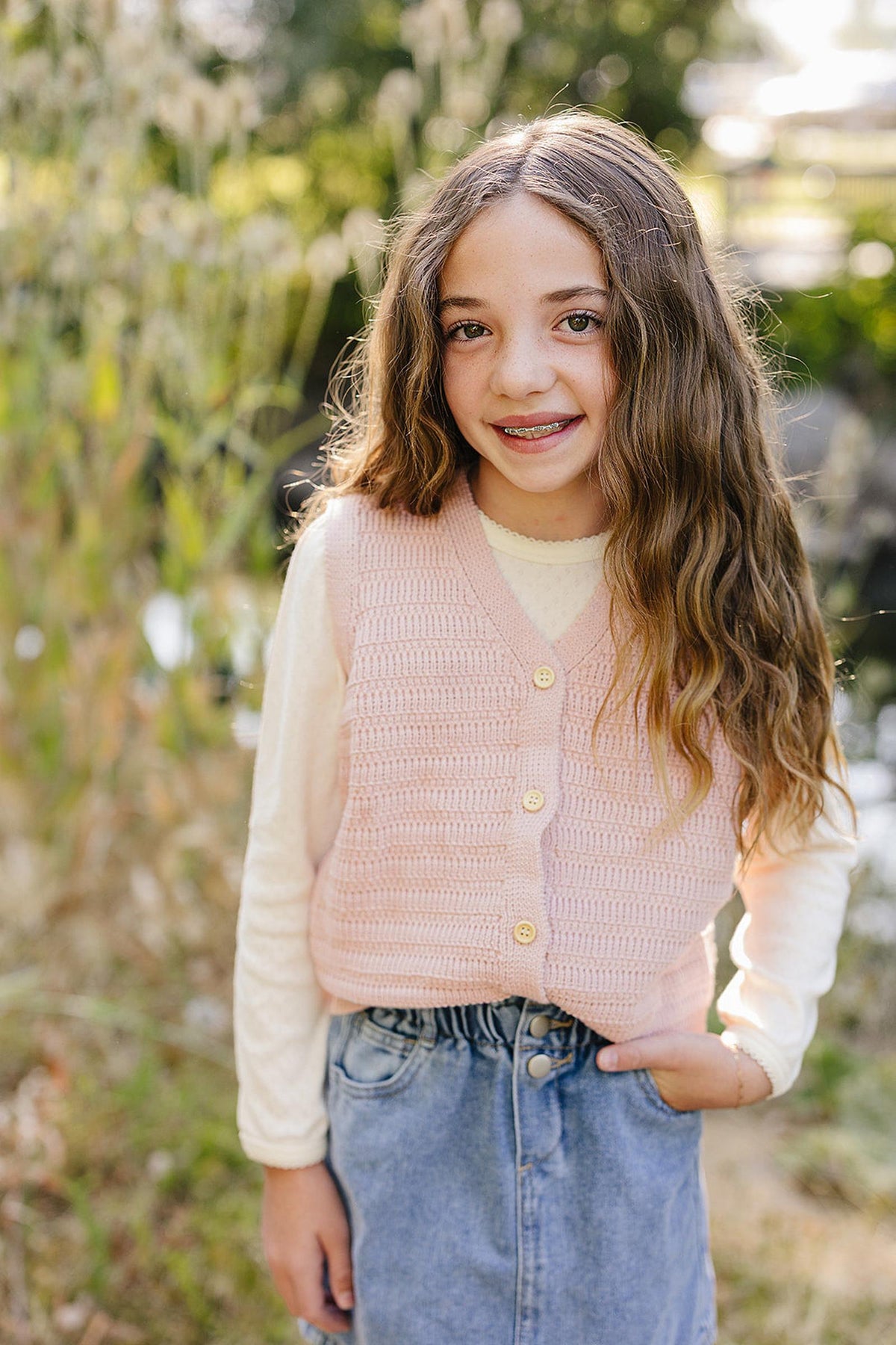 A child wearing a pink knit button-down vest over a cream shirt and denim skirt, standing outdoors.