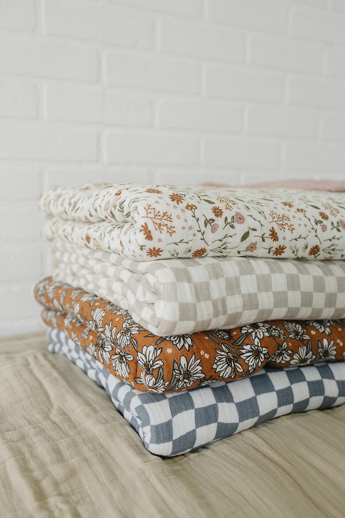 A stack of four folded muslin quilts in floral and checkered patterns on a neutral-colored surface.