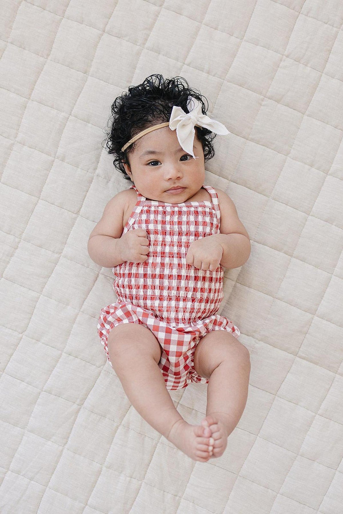 A baby with curly hair wears a red and white gingham romper, lying on a light quilted blanket.