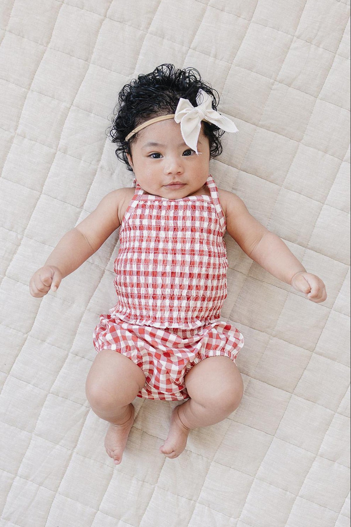 A baby girl lays on a quilted beige surface, wearing a red and white gingham romper with gentle ruching and.