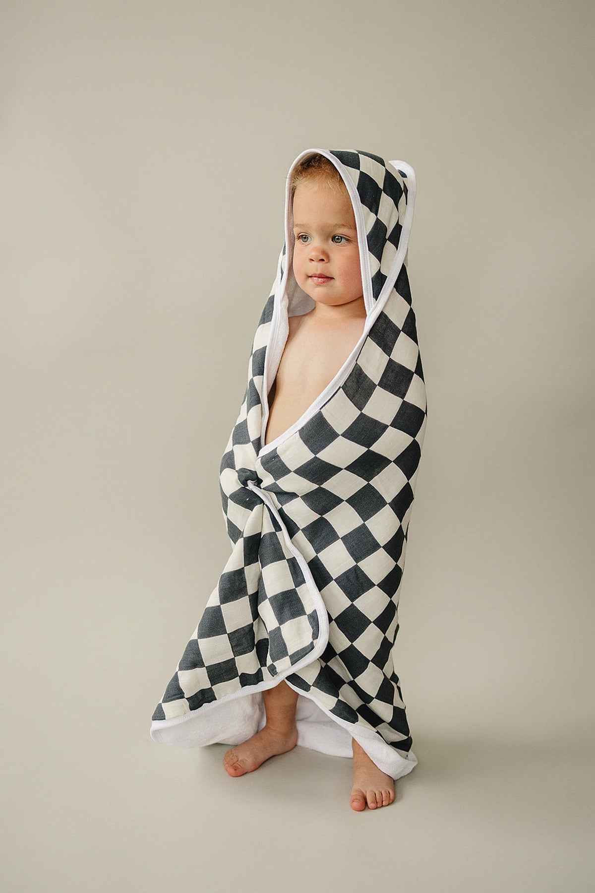 A baby wrapped in a charcoal checkered muslin hooded towel stands on a neutral background.