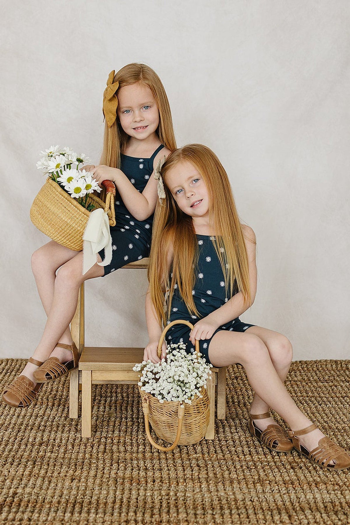 Two young girls in navy floral tank tops and shorts holding baskets of daisies, sitting on wooden stools.