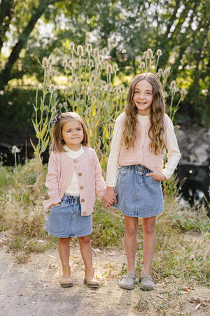 Two young girls stand outdoors, wearing pink knit tops and denim skirts, surrounded by tall plants and greenery.