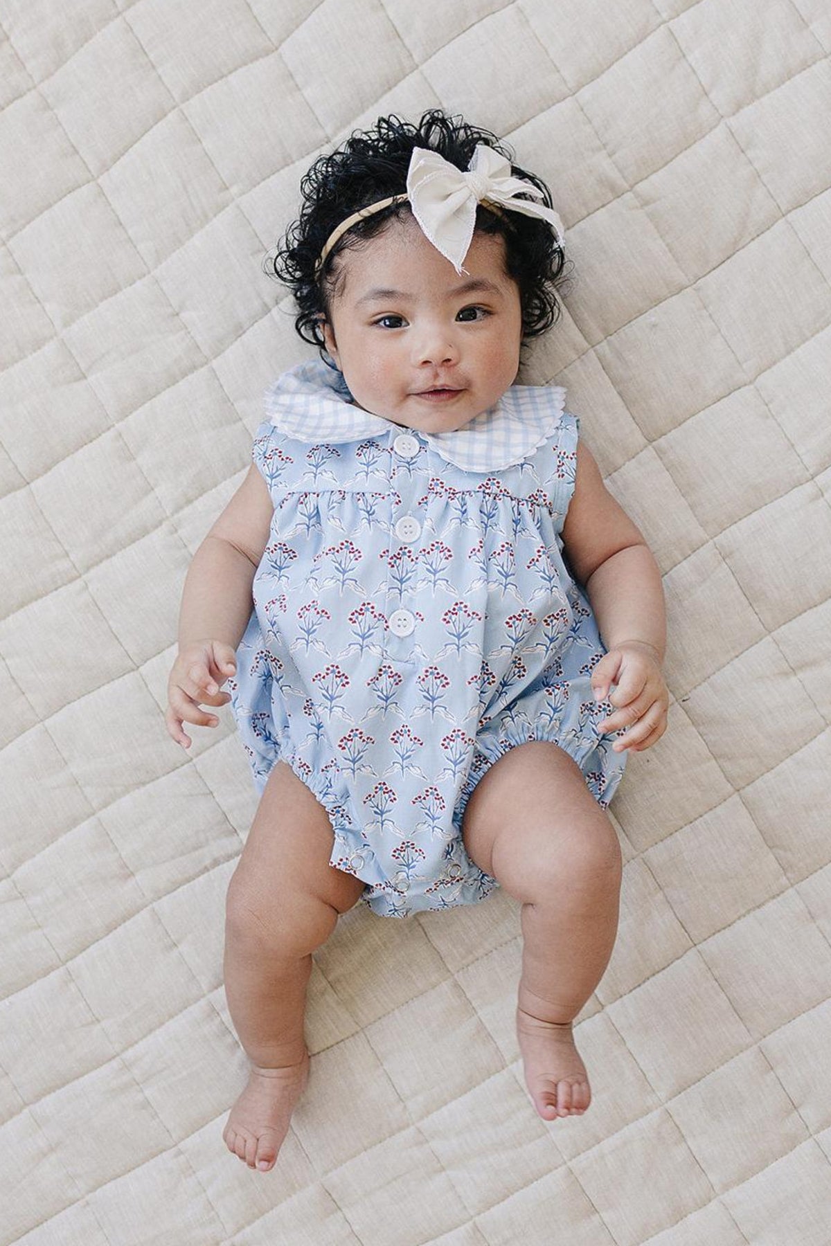 A baby girl lies on a textured beige blanket, wearing a light blue bubble romper adorned with delicate red flowers and a white collar.