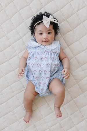 A baby girl lies on a textured beige blanket, wearing a light blue bubble romper adorned with delicate red flowers and a white collar.