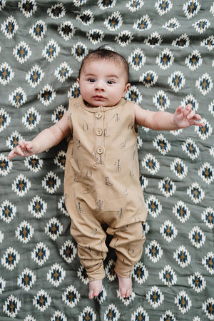 A baby lies on a patterned blanket, wearing a beige cotton romper with western-themed graphics, featuring buttons down the front.