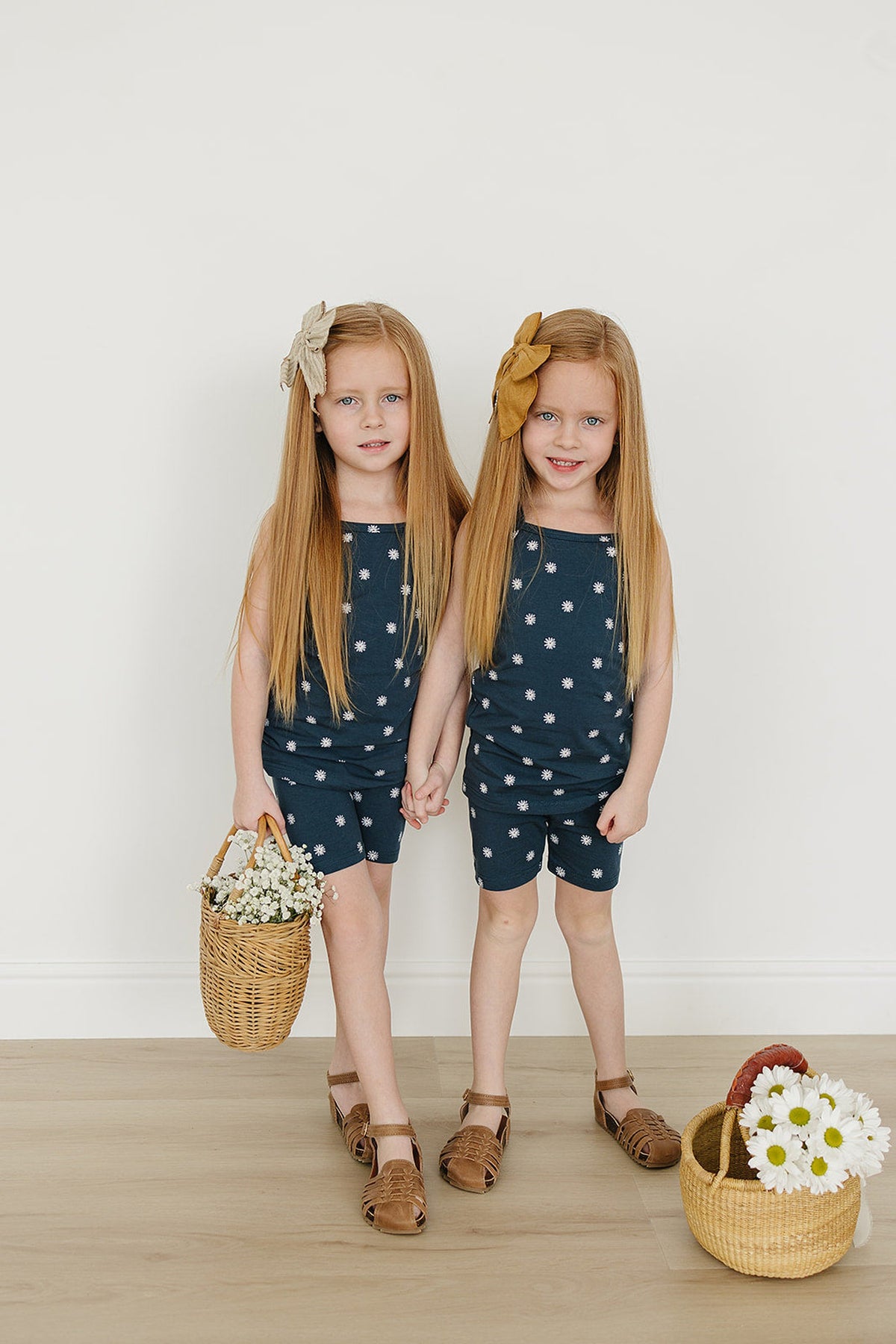 Two young girls wearing navy floral tank tops and biker shorts, holding hands with wicker baskets of white flowers.