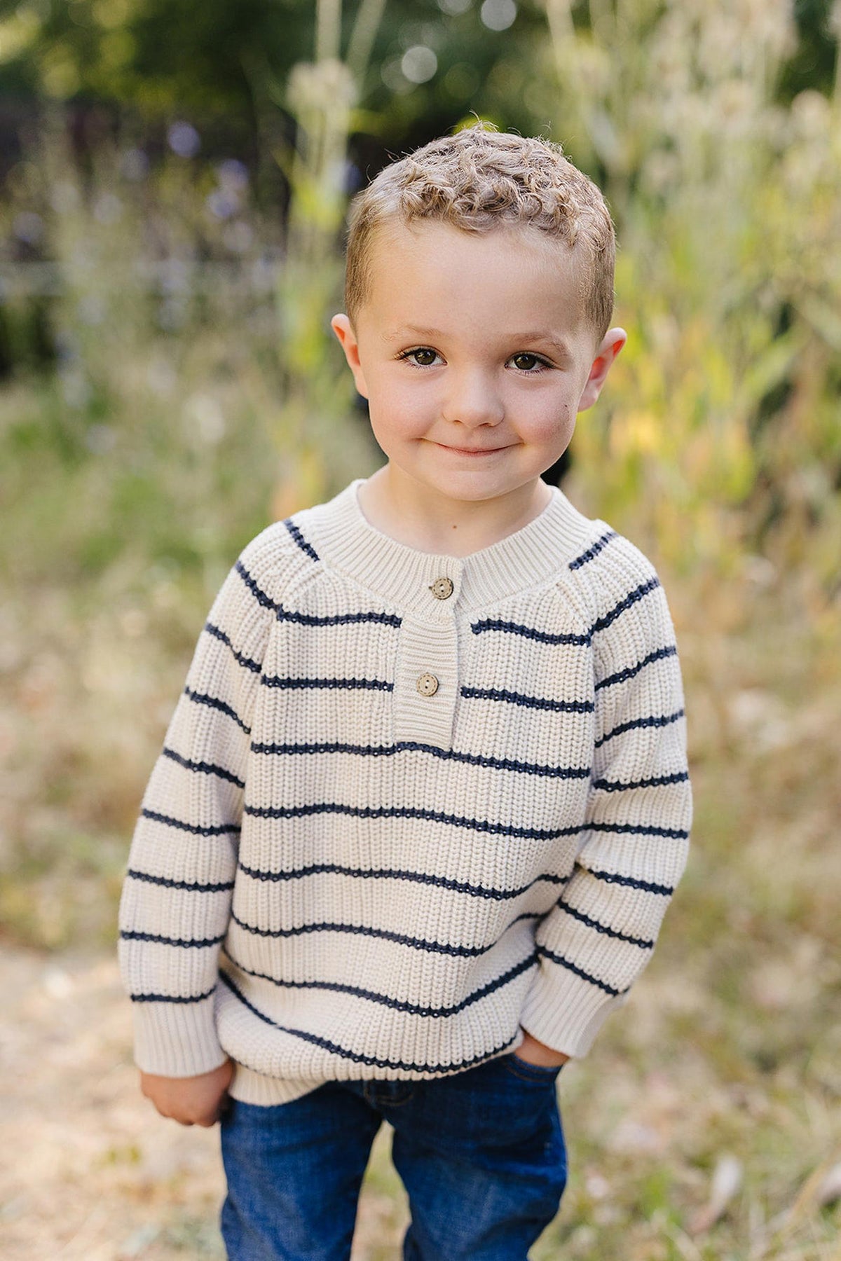 A child wearing a navy-striped knit sweater and jeans stands outdoors, smiling with a blurred natural background.