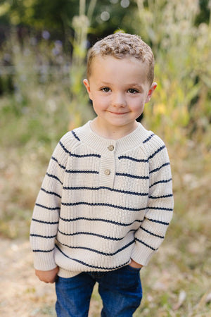 A child wearing a navy-striped knit sweater and jeans stands outdoors, smiling with a blurred natural background.