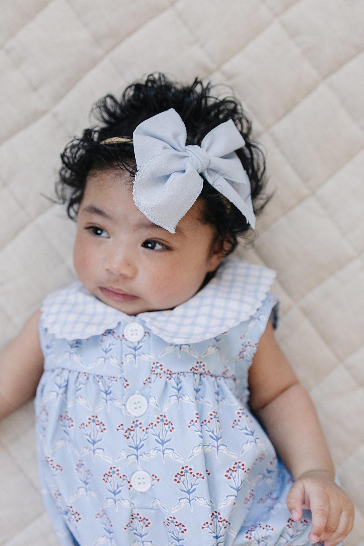 A baby girl with curly black hair wears a light blue floral romper featuring a white collar.
