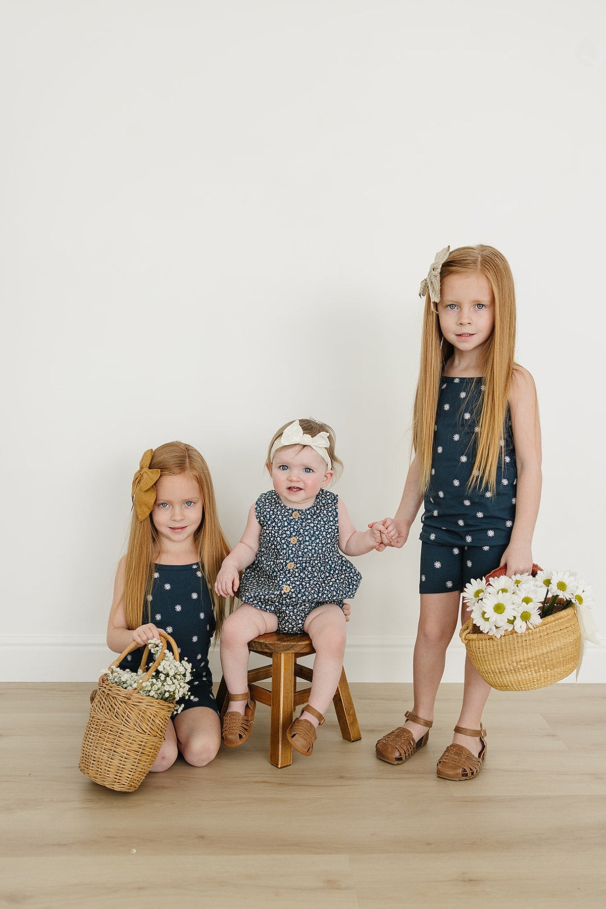 Three children in navy floral outfits, holding baskets with flowers, pose against a plain white wall.