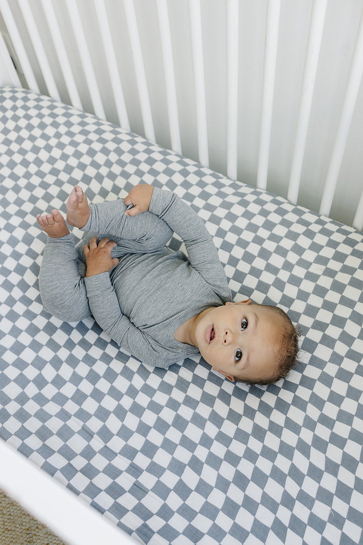 Baby lying on a wavy checkered muslin crib sheet in dusty blue, wearing a gray outfit.