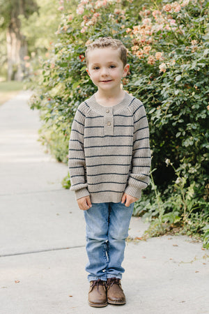 A young boy stands on a pathway surrounded by greenery and flowers, wearing a cozy brown and charcoal striped knit sweater, blue jeans, and brown shoes.
