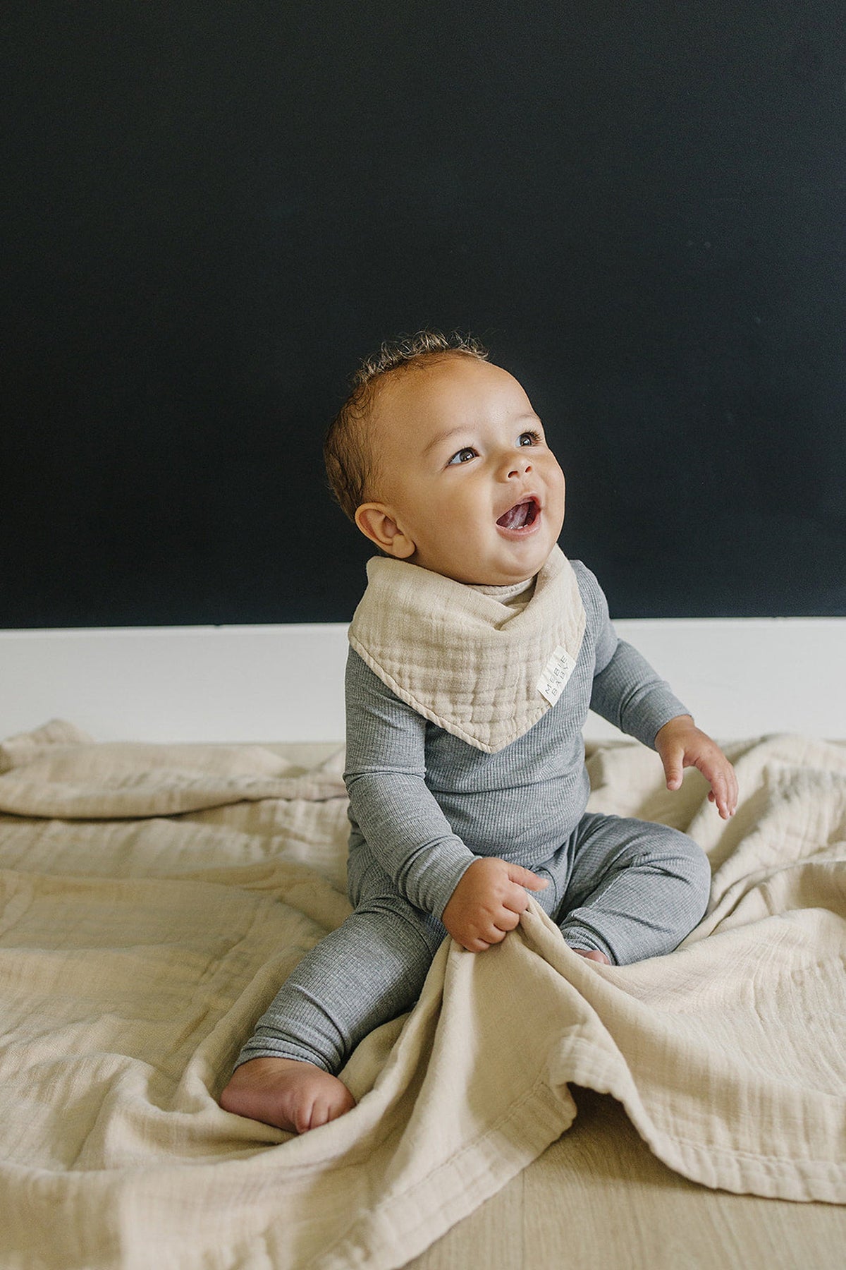 A baby in a gray outfit sits on an oatmeal muslin quilt, wearing a matching bib scarf, smiling.