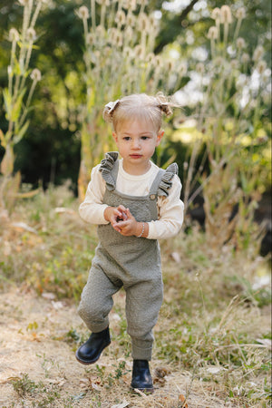 A toddler wearing green ruffle knit overalls and black boots stands in a grassy outdoor setting.