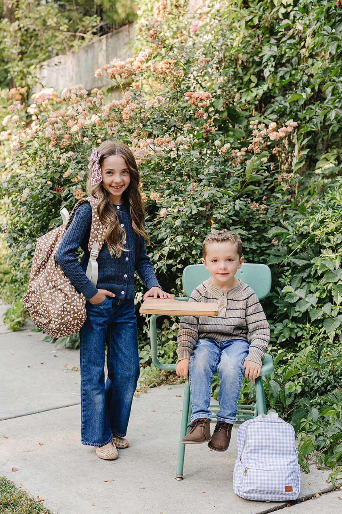 A girl in a navy cardigan and wide-leg jeans stands beside a boy seated on a green chair, both surrounded by lush greenery and blooming flowers, highlighting a playful outdoor.