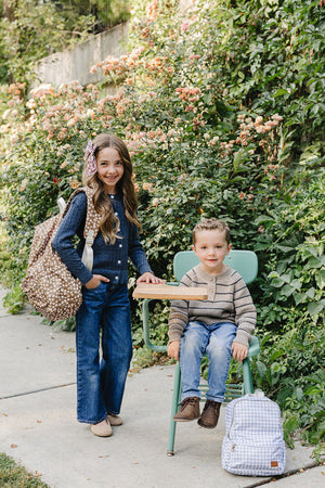 A girl in a navy cardigan and wide-leg jeans stands beside a boy seated on a green chair, both surrounded by lush greenery and blooming flowers, highlighting a playful outdoor.