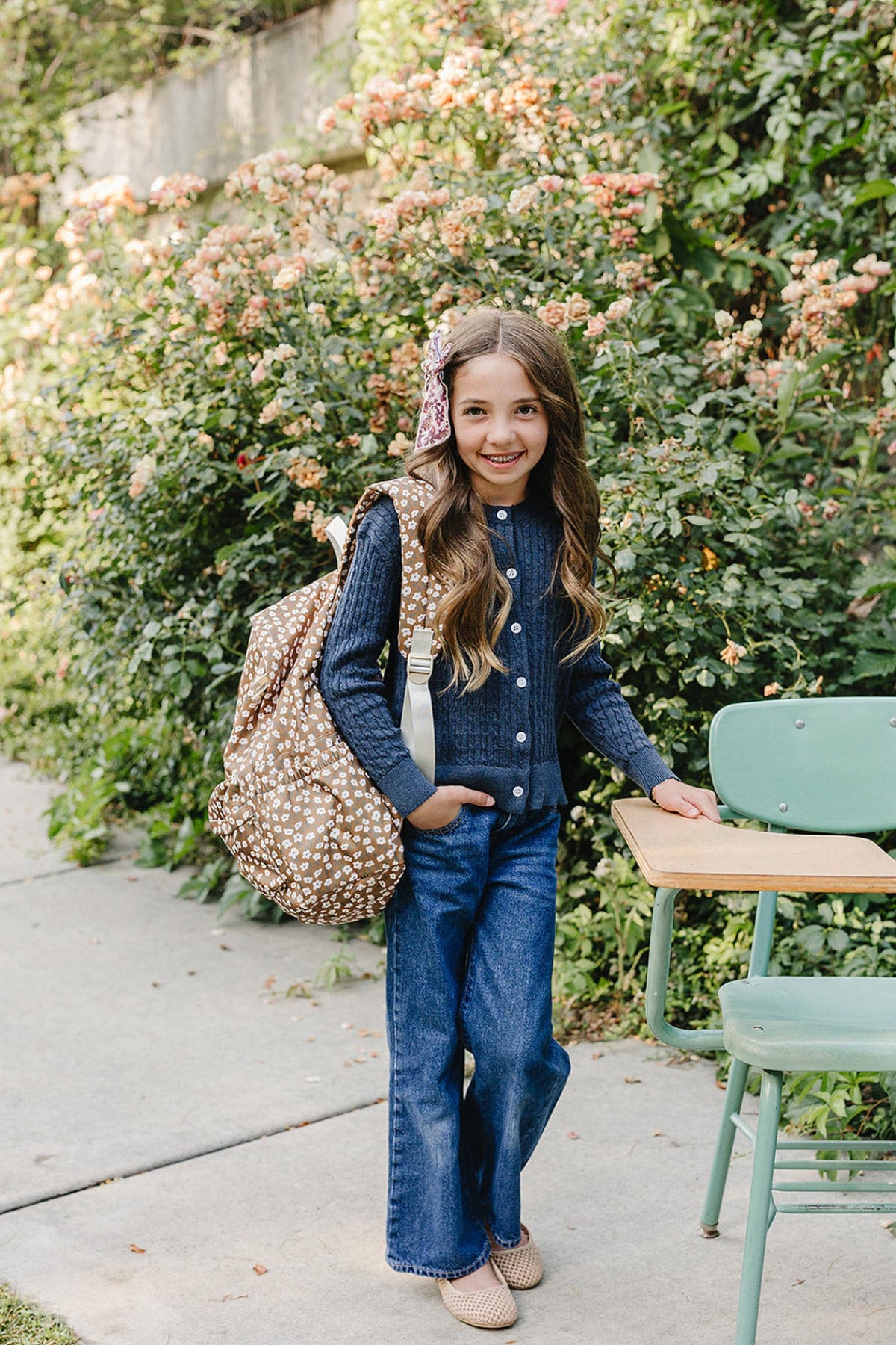 A young girl stands outdoors, smiling while wearing a navy cable knit cardigan and flared jeans. She carries a patterned backpack, surrounded by lush greenery and blooming flowers.