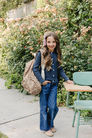 A young girl stands outdoors, smiling while wearing a navy cable knit cardigan and flared jeans. She carries a patterned backpack, surrounded by lush greenery and blooming flowers.