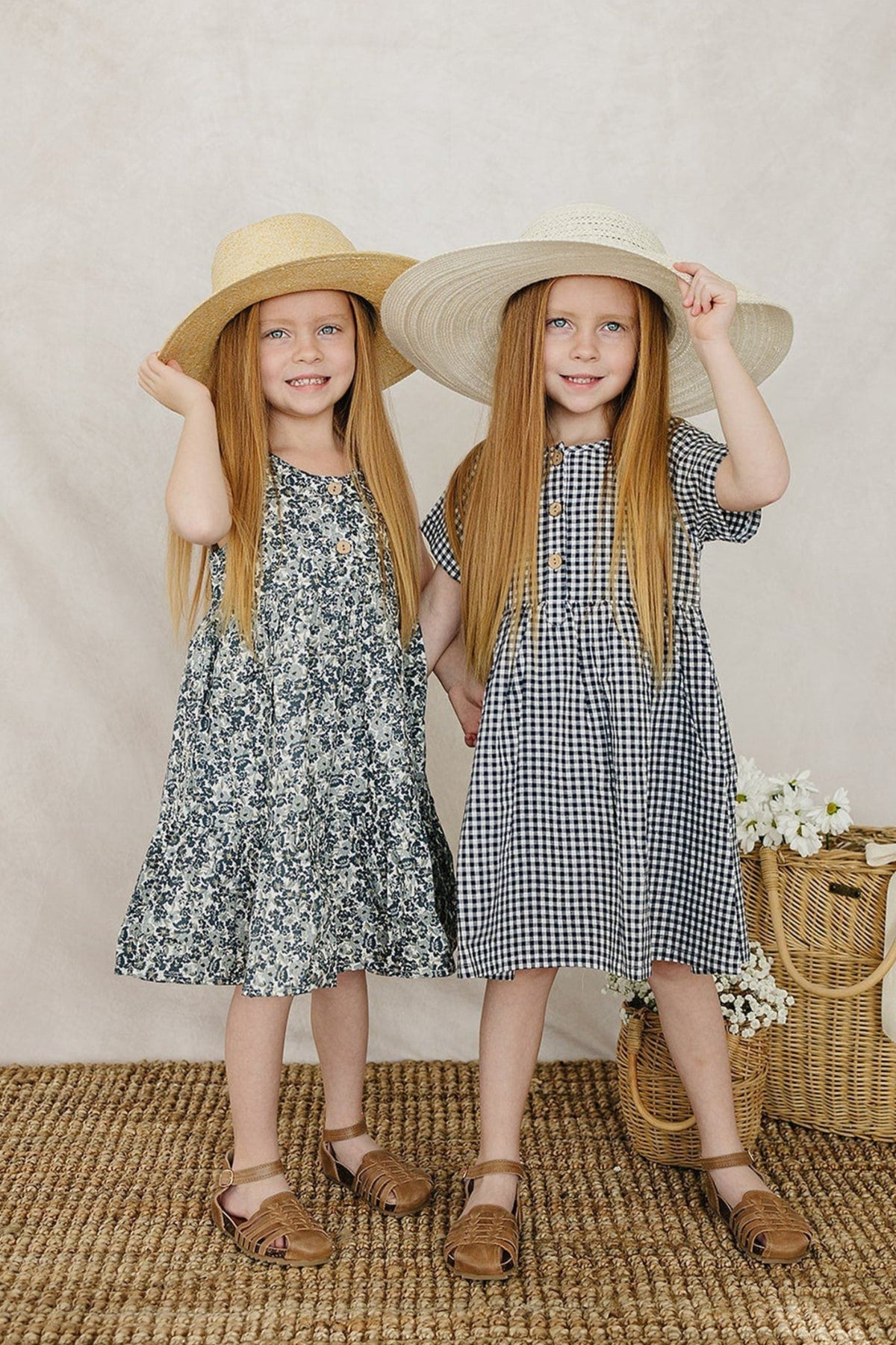 Two young girls wearing floral and gingham dresses stand side by side, each holding a wide-brimmed straw hat. The background features a soft, neutral fabric, enhancing the playful summer vibe.