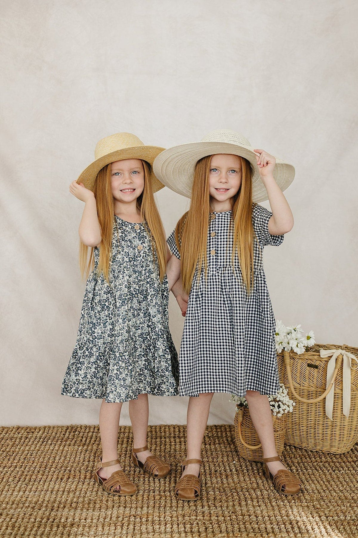 Two young girls stand side by side, wearing summer dresses—one floral and one navy gingham—holding wide-brimmed hats.