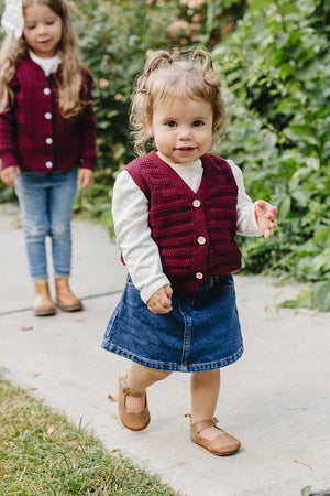 A young girl walks on a path wearing a burgundy knit cotton vest over a white shirt and denim skirt. She smiles, showcasing the vests soft texture and classic style.